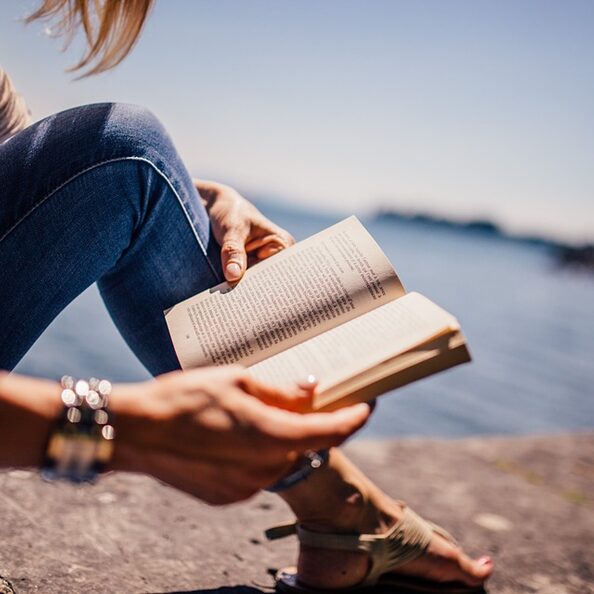 Auf dem Foto ist eine Frau zu erkennen, die ein Buch  liest. Sie sitzt auf einer Mauer, im Hintergrund erkennt man einen See oder auch das Meer.