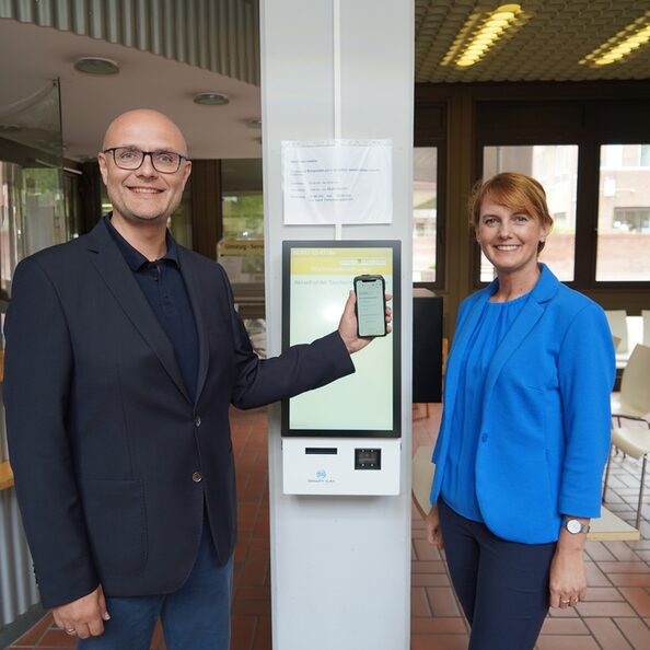 Gudrun van Cleef (r.) und Norbert Sester vor dem Bondrucker des Bürgeramts mit Smartphone.