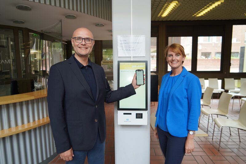 Gudrun van Cleef (r.) und Norbert Sester vor dem Bondrucker des Bürgeramts mit Smartphone.